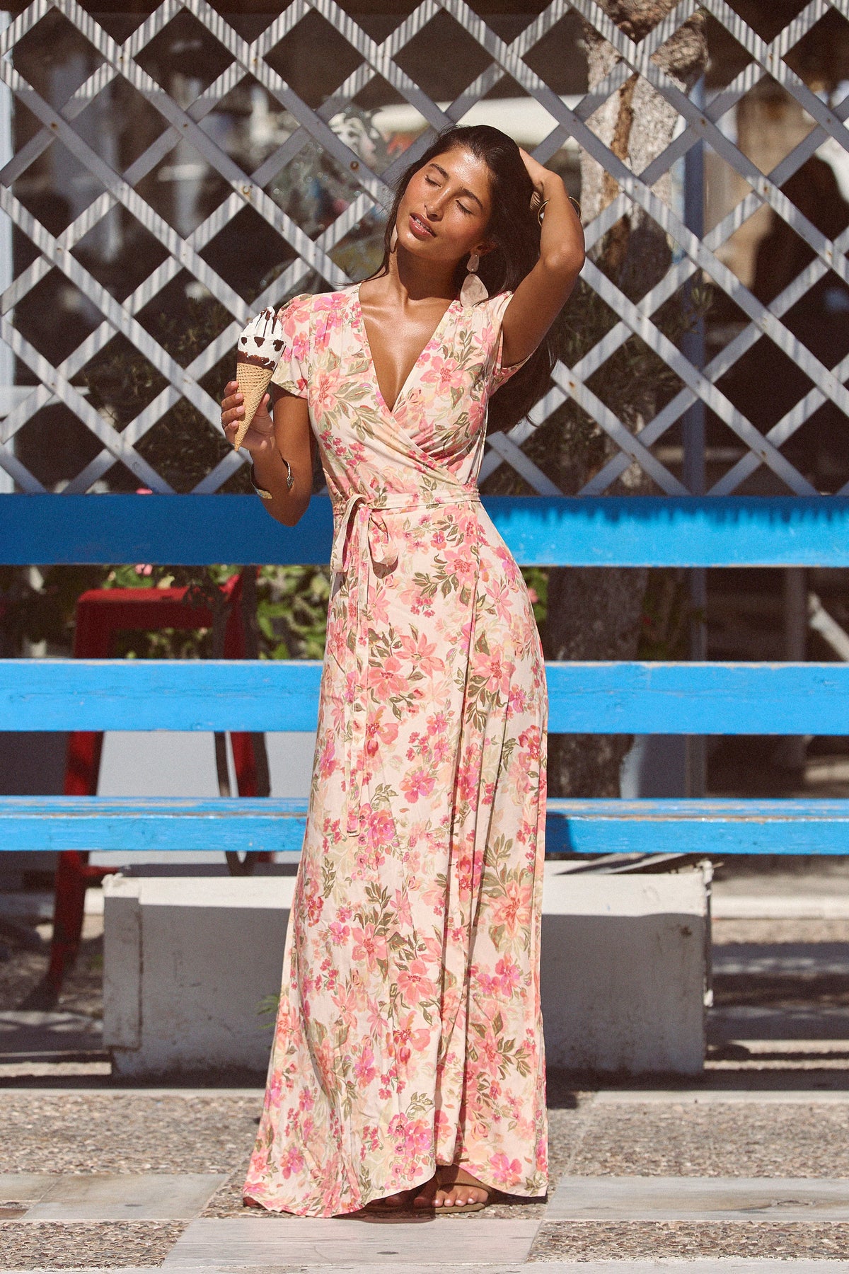 A woman stands outdoors in front of a blue bench and lattice fence, wearing a long floral wrap maxi dress with a V-neck and tie waist. She holds an ice cream cone.