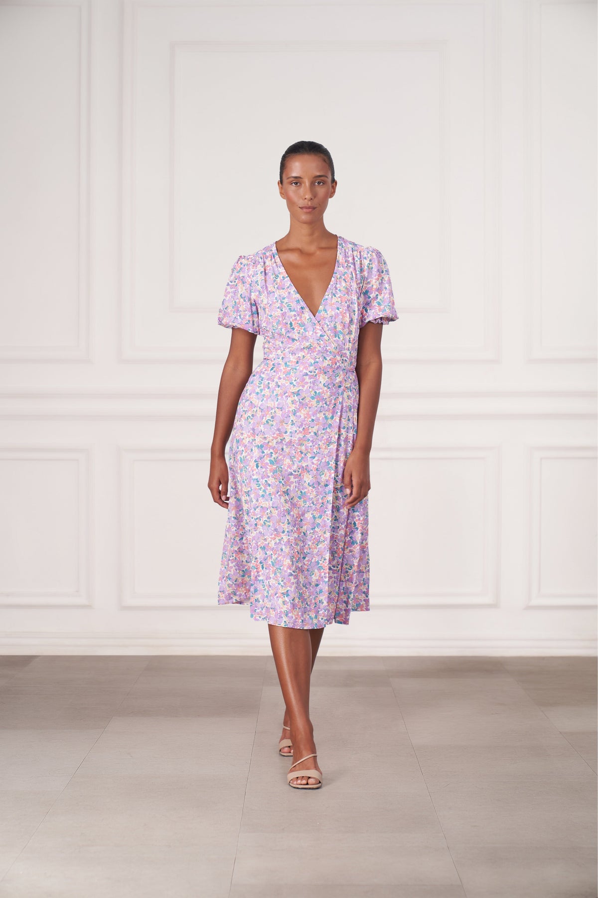 A woman modeling a short-sleeve wrap-style midi dress with a light purple, pink, and blue floral print, standing in front of a white paneled wall.