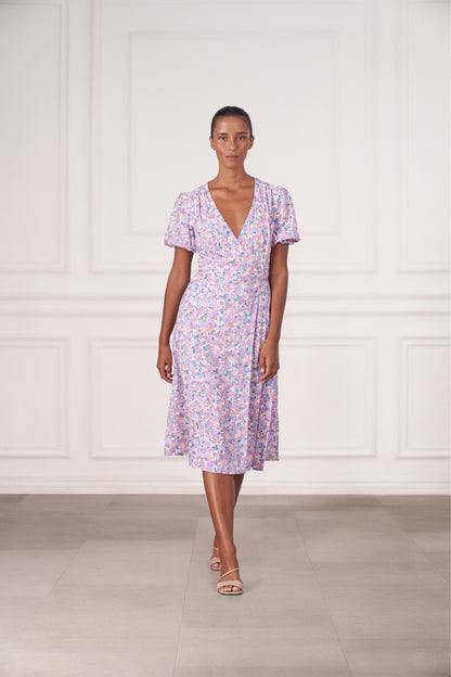 A woman modeling a short-sleeve wrap-style midi dress with a light purple, pink, and blue floral print, standing in front of a white paneled wall.
