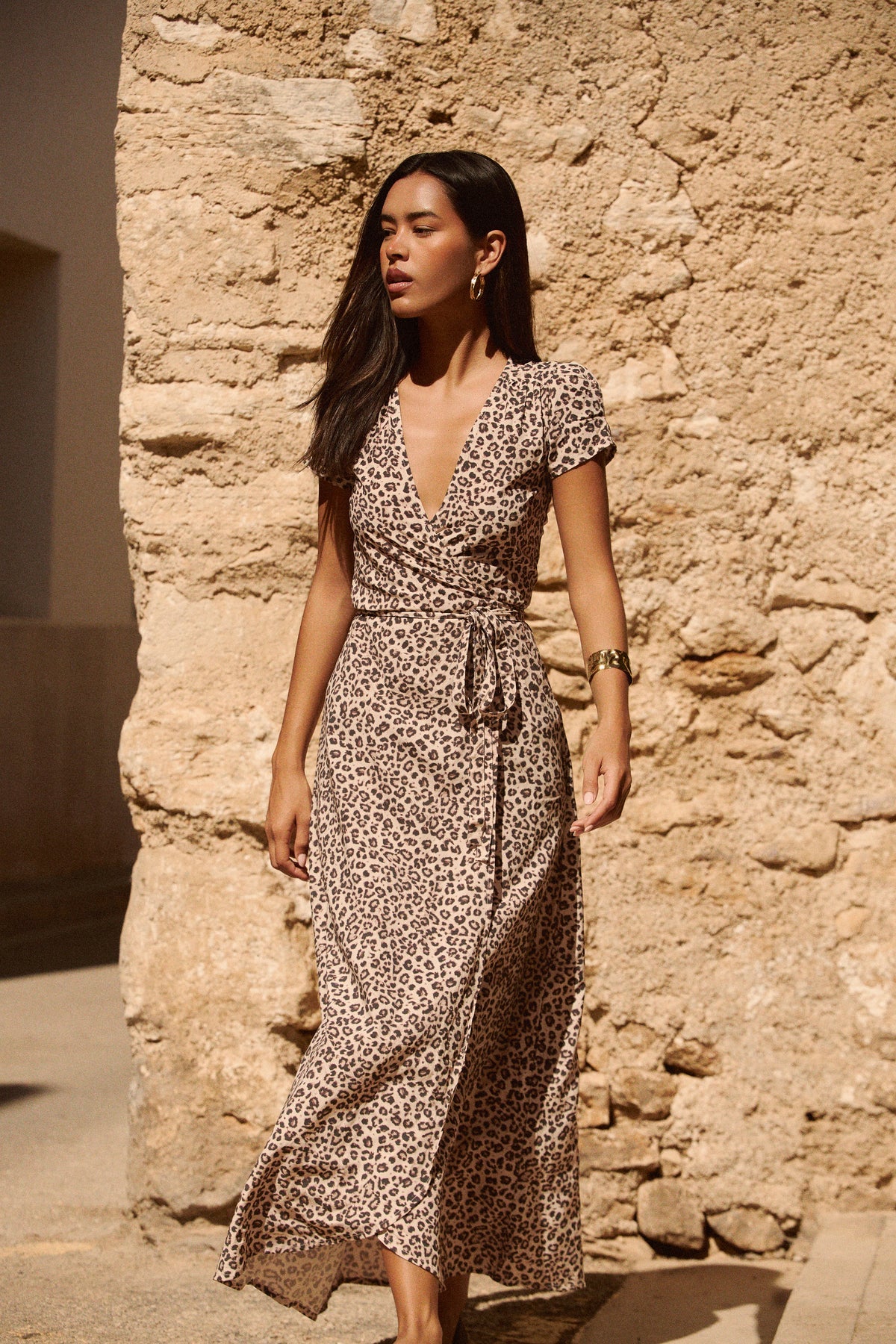 A woman wearing a leopard-print maxi wrap dress with short flutter sleeves and a tie waist, standing outdoors against a rustic beige stone wall.