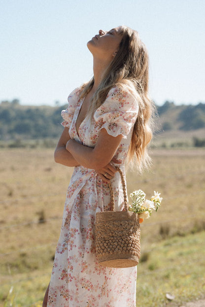 A woman in a white floral puff-sleeve maxi dress stands outdoors in a field, holding a woven straw tote bag with white flowers. She is looking upward under a clear blue sky.