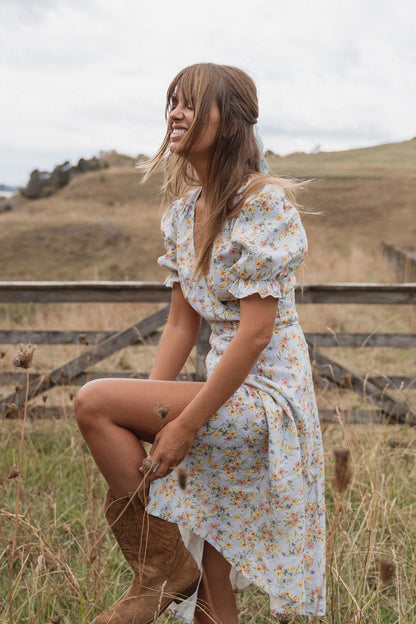 A woman wearing a white floral puff-sleeve dress and brown cowboy boots sits in a grassy field with a wooden fence and rolling hills in the background.