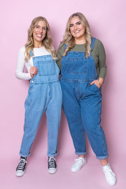Two women wearing blue denim overalls against a pink background